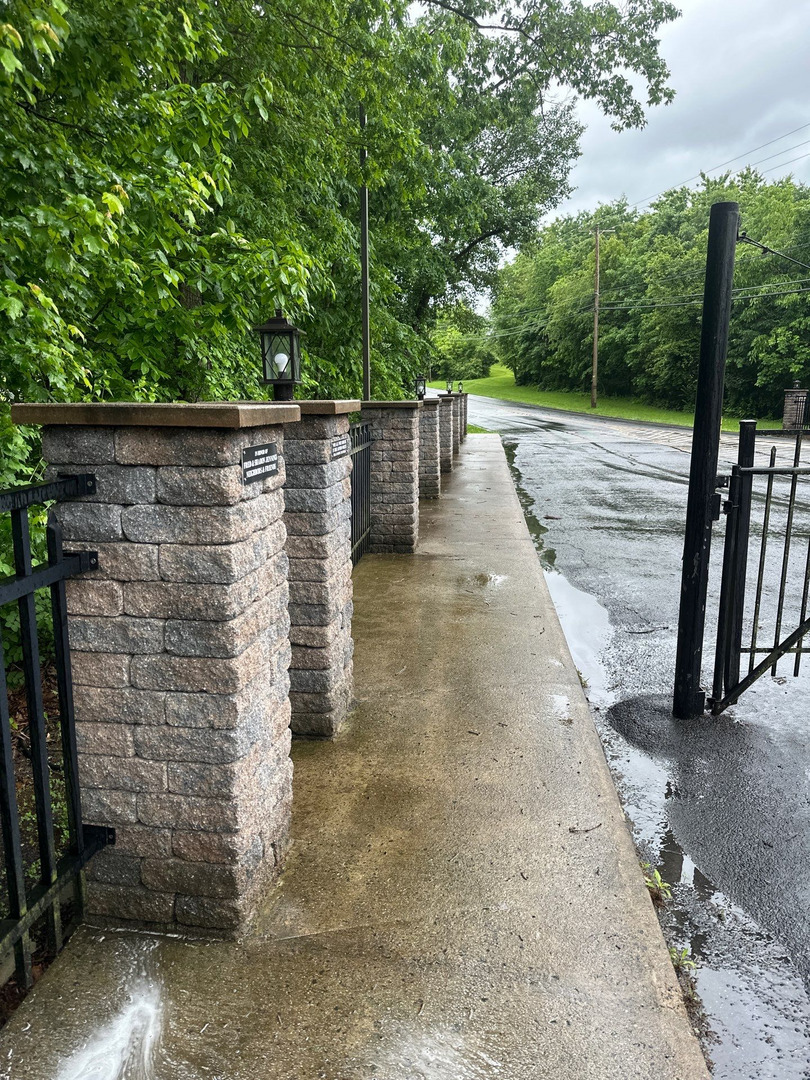 cleaning and rinsing a concrete surface near a stone wall and black metal gate surrounded by greenery